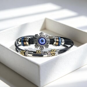 May include: Two black leather bracelets with silver and gold-toned metal accents. One bracelet features a flower-shaped charm with a blue eye design. The bracelets are displayed in a white box.