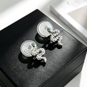 May include: A pair of silver-toned snake-shaped clip-on earrings featuring a textured, coiled body design and a small, vibrant green stone set as the eye. The earrings are attached to clear, circular silicone pads for comfortable wear.