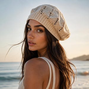 Op de afbeelding: Een beige gebreide baret met een ruitpatroon. De baret wordt op het hoofd gedragen. De persoon draagt een crèmekleurige top met spaghettibandjes. De achtergrond is een strandscène met de oceaan en de lucht.