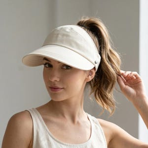 Op de afbeelding: Een crèmekleurige vizierhoed met een gebogen rand. De hoed heeft een gestructureerd ontwerp en een band aan de achterkant. De vizier wordt gedragen door een persoon met haar in een paardenstaart.