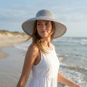 Puede incluir: Un sombrero de sol de ala ancha con un patrón tejido gris y blanco. El sombrero lo lleva una persona en una playa, con el océano y el cielo de fondo. El sombrero proporciona sombra y estilo.