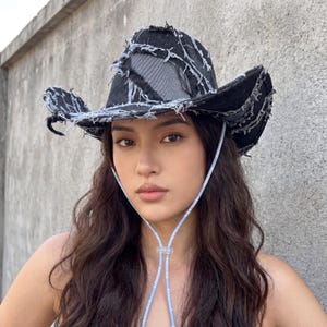 Può includere: Un cappello da cowboy in denim nero con bordi usurati e un pannello grigio. Il cappello presenta cuciture bianche e un sottogola azzurro. La tesa del cappello è leggermente curva e lo stile generale è audace e moderno.