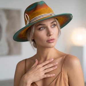 Może przedstawiać: Kapelusz typu fedora z szerokim rondem, ozdobiony ręcznie malowanym gradientem w odcieniach turkusu, pomarańczu i żółci. Kapelusz posiada ozdobną biało-czarną wzorzystą taśmę z małą pętelką z boku oraz widoczne białe przeszycia na krawędzi ronda.