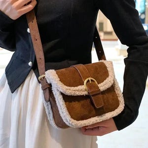 May include: A brown suede crossbody bag with a cream-colored, fluffy trim. The bag features a flap closure with a gold-colored buckle and a brown adjustable strap. The bag is held by a person wearing a black jacket and a white skirt.