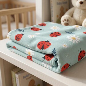 Op de afbeelding: Gevouwen lichtblauwe babydeken met een herhalend patroon van rode lieveheersbeestjes en witte madeliefjes. De deken is gemaakt van zacht materiaal en is netjes op een witte plank gevouwen. Onderdeel van een babyproductcollectie.