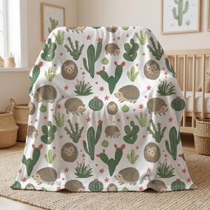 May include: A white blanket featuring a repeating pattern of brown hedgehogs, green cacti, and red star-shaped flowers. The blanket is draped over a chair, showcasing the design. The blanket is made of a soft material.