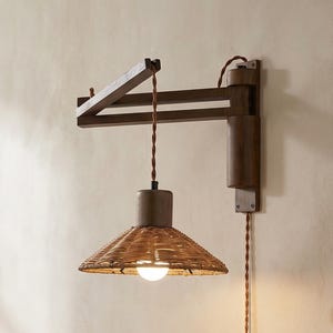 May include: A wall-mounted lamp with a dark brown wooden arm and a woven rattan lampshade. The lamp has a brown cord and a cylindrical metal base. The light is on, illuminating the woven shade. The lamp is designed for indoor use.
