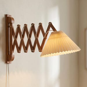May include: A wall-mounted lamp with an extendable wooden arm and a pleated lampshade. The lamp is made of dark wood and has a cream-coloured lampshade. The lamp is plugged into a wall socket.