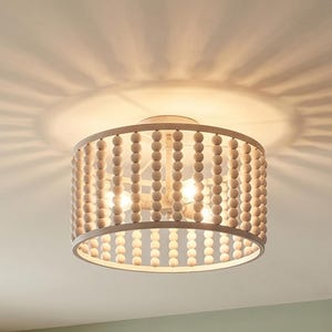 May include: A flush mount ceiling light fixture with a drum shade. The shade is made of light-colored wooden beads. The fixture has a circular frame and three light bulbs. The light casts a pattern on the ceiling.