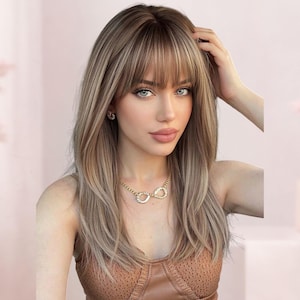 Op de afbeelding: Een lange, gelaagde pruik met een lichtbruine en asblonde balayage kleurenpalet met een rechte, warrige pony die het voorhoofd omlijst. Het haar heeft een gladde, slanke textuur met subtiele highlights en valt tot voorbij de schouders.