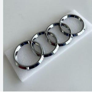 May include: A chrome-finished emblem featuring the iconic four interlocking rings, a symbol of automotive excellence. The emblem is presented on a white foam surface, showcasing its polished, reflective surface.
