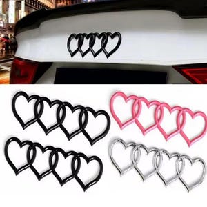 May include: Heart-shaped car emblems in black, pink, and silver. The emblems are designed to resemble the Audi logo, with four interlocking hearts. One emblem is attached to a white car.