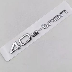 May include: A chrome-finished emblem with the text "40 e-tron" on a white rectangular backing. The numbers and letters are in a bold, sans-serif font, with a slight 3D effect. The emblem is likely for a vehicle.