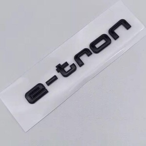 May include: Black "e-tron" emblem on a white background. The letters are bold and three-dimensional, with a modern, geometric font. The emblem is likely a car badge or sticker.
