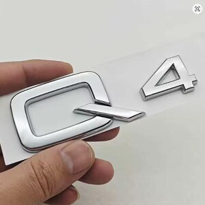 Può includere: Emblema cromato Q4, probabilmente per un veicolo. La 'Q' è una lettera stilizzata con un'estensione rettangolare. Il numero '4' è separato. Entrambi gli elementi sono lucidi e su sfondo bianco.