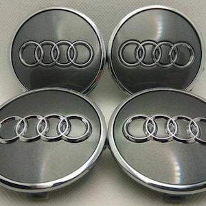 May include: Four gray and silver car wheel center caps with the Audi logo. Each cap features a gray center with the Audi rings logo in silver, surrounded by a silver rim. The caps are circular.