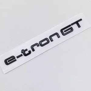 Może przedstawiać: Czarny emblemat e-tron GT na białej prostokątnej naklejce. Litery są pogrubione i bezszeryfowe, z błyszczącym wykończeniem. Naklejka jest lekko pochylona, o czystej, minimalistycznej estetyce.