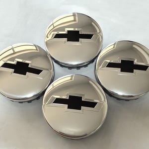 May include: Four chrome hubcaps with a black Chevrolet bow tie logo. The hubcaps are round and have a shiny, reflective surface. The black logo is centered on each hubcap.