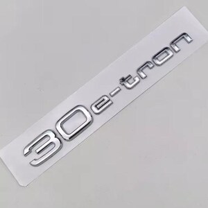 May include: Silver 30 e-tron emblem on a white rectangular backing. The emblem features the number "30" followed by "e-tron" in a modern font. The emblem is likely for a vehicle.