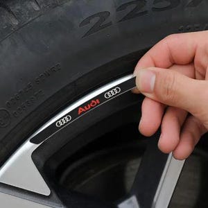 May include: A close-up of a black and silver car wheel with a tire. A hand is holding a black and white Audi sticker with red lettering. The Audi logo is visible on the sticker.
