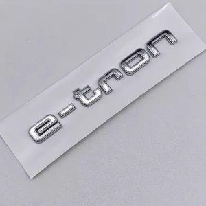 Könnte beinhalten: Ein verchromtes "e-tron"-Emblem auf einem weißen rechteckigen Träger. Die Buchstaben sind in einer modernen, serifenlosen Schriftart mit reflektierendem, metallischem Finish gehalten. Das Emblem ist wahrscheinlich für den Automobilbereich bestimmt, möglicherweise ein Autoabzeichen.