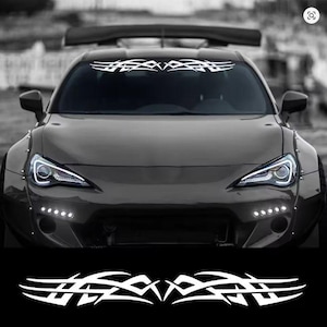 Puede incluir: Un coche deportivo negro con una calcomanía de diseño tribal blanco en la parte superior del parabrisas. El coche tiene faros brillantes y un alerón. Se muestra una calcomanía a juego debajo.