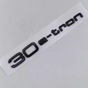 May include: Black 3D emblem with the text "30 e-tron" on a white rectangular background. The numbers and letters are in a bold, sans-serif font. The emblem appears to be made of a glossy material.