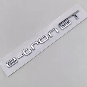 May include: A chrome-colored "e-tron GT" emblem, affixed to a white backing. The letters are in a modern, sans-serif font, with a reflective, metallic finish. The emblem is likely for a vehicle, showcasing the model name.