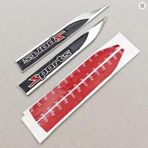 Puede incluir: Dos emblemas de coche "Sports" plateados y negros con detalles en rojo. Los emblemas son alargados y tienen respaldo adhesivo para una fácil aplicación. La imagen también muestra el respaldo adhesivo rojo.