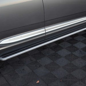 May include: A side step for a vehicle, featuring a silver and black design. The step is mounted along the lower side of the car, with the word "QUATTRO" visible on the side. The vehicle's body is a dark gray color.