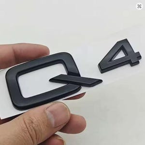 May include: Black Q4 car emblem with a glossy finish. The emblem features the letter "Q" and the number "4" in a bold, sans-serif font. The accessory is held against a white background. A car styling accessory.