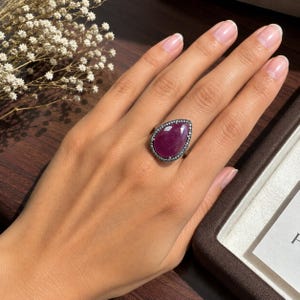 May include: A teardrop-shaped ruby ring featuring a large, deep pinkish-red gemstone set in a dark metal band. The central stone is surrounded by a delicate halo of tiny, sparkling white accent stones, creating a refined and elegant appearance.