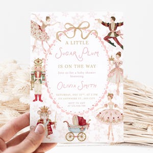 Könnte beinhalten: Eine rechteckige Babyparty-Einladungskarte mit weißem Hintergrund und rosa Schneeflockenmustern. Die Illustrationen zeigen einen Nussknacker, Ballerinas, einen Mäusekönig und einen Kinderwagen. Text: A LITTLE SUGAR PLUM IS ON THE WAY.