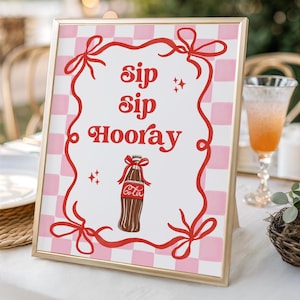 Könnte beinhalten: Ein rechteckiges Schild mit goldenem Rahmen und einem rosa-weiß karierten Hintergrund. Der rote Schriftzug "sip sip Hooray" ist von einer roten Schleifenbordüre umgeben, mit einer zentralen Abbildung einer braunen Cola-Flasche.