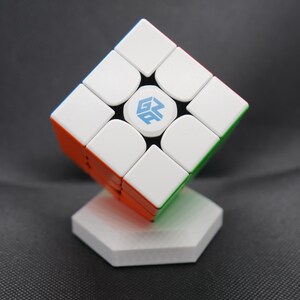 May include: A white, orange, green, and blue speed cube puzzle, displayed on a white hexagonal stand. The cube has a logo in the center. The cube is a classic brain teaser.