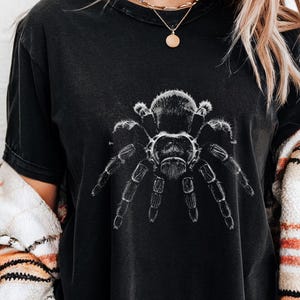 以下が含まれることがあります： 黒いTシャツ。白いタランチュラの詳細なイラストが描かれています。Tシャツはヴィンテージ風の外観です。「Black」の文字が右下に白でプリントされています。衣料品です。