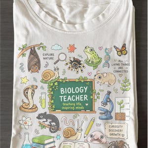 May include: White t-shirt featuring a colorful biology-themed design. The graphic includes various animals, plants, and scientific elements, with the text "BIOLOGY TEACHER" and "teaching life, inspiring minds".