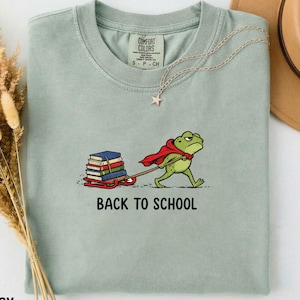 以下が含まれることがあります： 薄い緑色のTシャツ。マントを着たカエルのグラフィックが、本を載せたそりを引いています。「BACK TO SCHOOL」の文字が下にあります。星のペンダントが付いたゴールドのネックレスがシャツの上にあります。