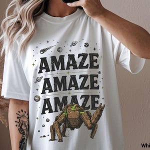 May include: White t-shirt with the word "AMAZE" repeated three times in a bold, black font with green accents. The design includes space-themed graphics like planets, stars, and a robotic creature. The shirt is a casual, oversized style.