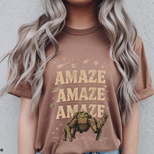 May include: Espresso-colored t-shirt with the word "AMAZE" repeated three times in a vintage font. The shirt features a graphic of a creature with a raised arm, surrounded by planets and stars.