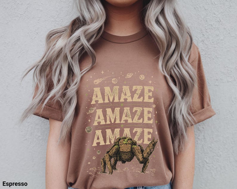 May include: Espresso-colored t-shirt with the word "AMAZE" repeated three times in a vintage font. The shirt features a graphic of a creature with a raised arm, surrounded by planets and stars.