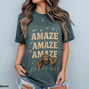 May include: Blue spruce t-shirt with the word "AMAZE" repeated three times in a tan font. A brown and green creature illustration is below the text. The shirt has a crew neck and short sleeves.