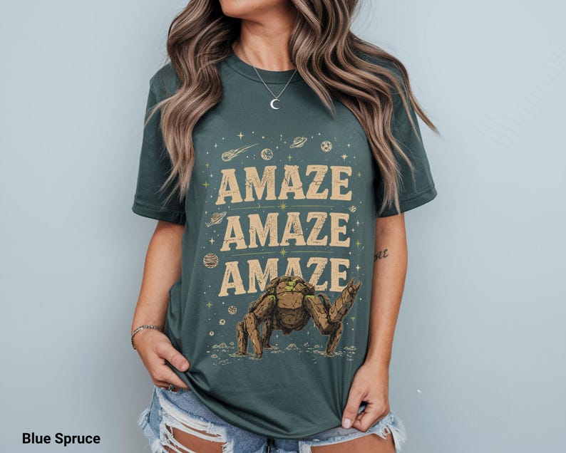 May include: Blue spruce t-shirt with the word "AMAZE" repeated three times in a tan font. A brown and green creature illustration is below the text. The shirt has a crew neck and short sleeves.