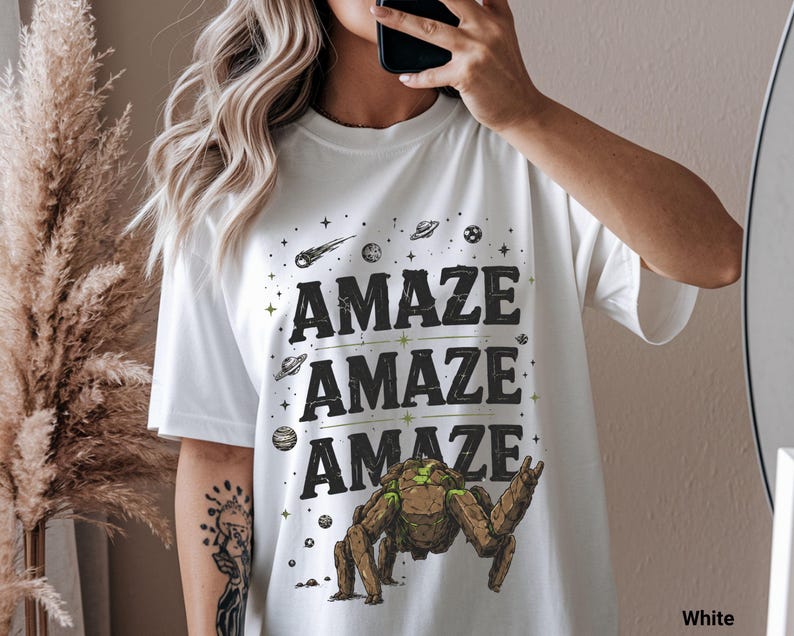 May include: White t-shirt with the word "AMAZE" repeated three times in a bold, black font with green accents. The design includes space-themed graphics like planets, stars, and a robotic creature. The shirt is a casual, oversized style.