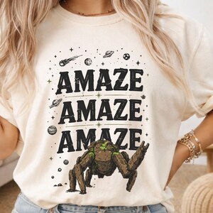 May include: Ivory-colored t-shirt with the word "AMAZE" repeated three times in black, with a space-themed graphic. The graphic includes a robot-like creature, planets, stars, and a comet.