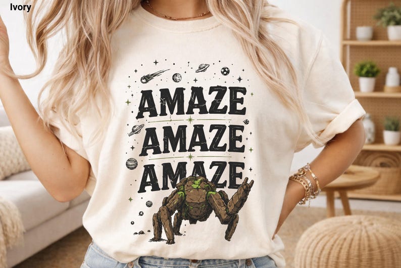 May include: Ivory-colored t-shirt with the word "AMAZE" repeated three times in black, with a space-themed graphic. The graphic includes a robot-like creature, planets, stars, and a comet.