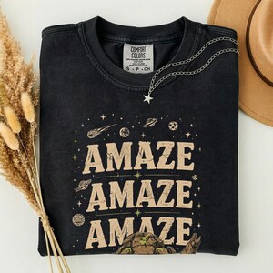May include: Black t-shirt with the word "AMAZE" in a tan font, repeated three times with space-themed illustrations. A silver necklace with a star pendant is draped on the shirt. The shirt has a Comfort Colors tag.