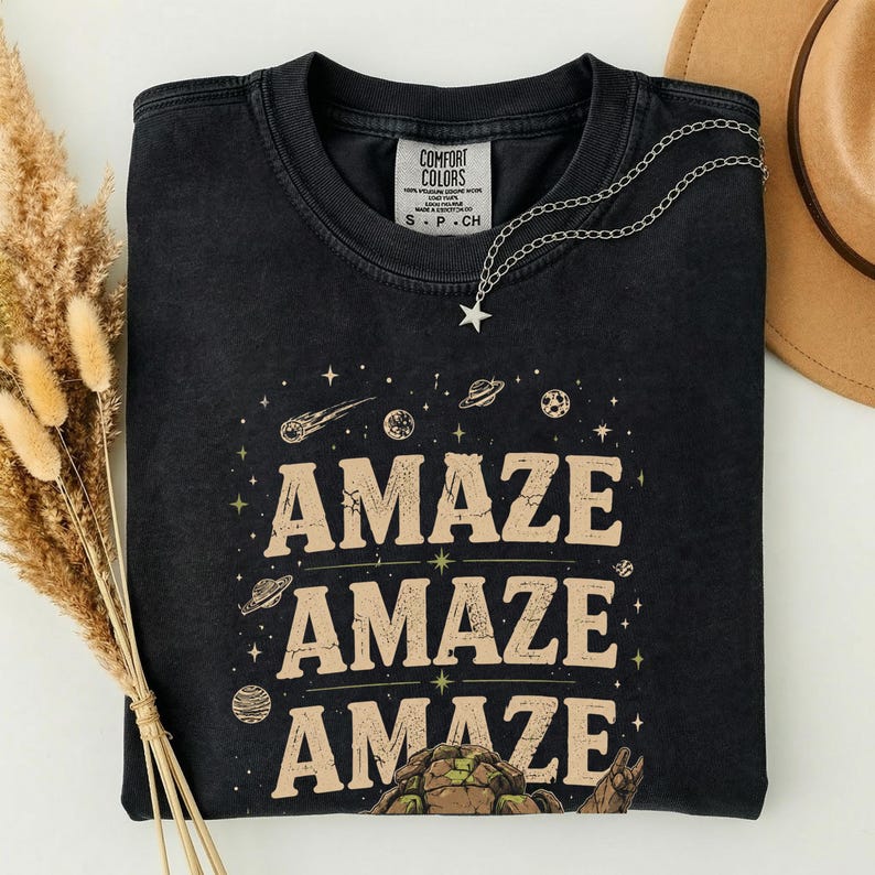 May include: Black t-shirt with the word "AMAZE" in a tan font, repeated three times with space-themed illustrations. A silver necklace with a star pendant is draped on the shirt. The shirt has a Comfort Colors tag.