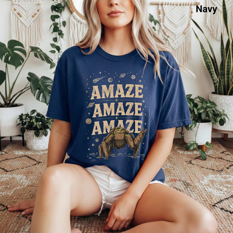 May include: Navy blue t-shirt with the word "AMAZE" repeated three times in a tan font. A cartoon creature is below the text. The shirt has a crew neck and short sleeves. The word "Navy" is in the top right corner.