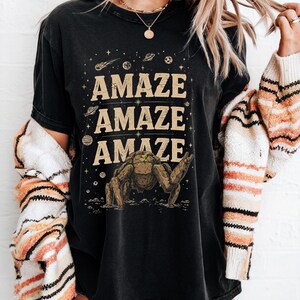 May include: Black t-shirt with the word "AMAZE" repeated three times in a tan font. A brown creature graphic is below the text, with planets and stars in the background. The word "Black" is at the bottom right.